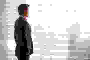 Business man stands looking out out a blurred window with hands in pockets