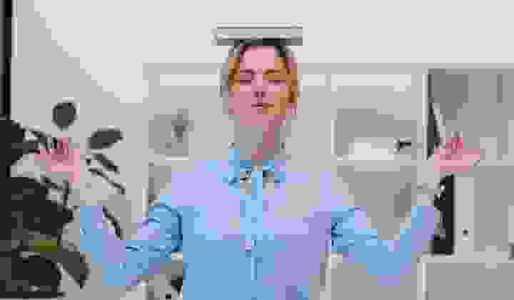 Woman standing balancing a book on her head with hands out and eyes closed