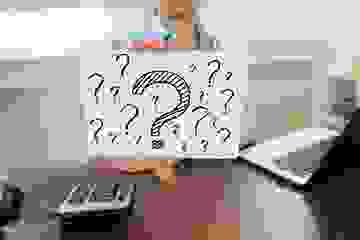 Woman at her desk with a calculator and laptop holding up a sign with lots of question marks