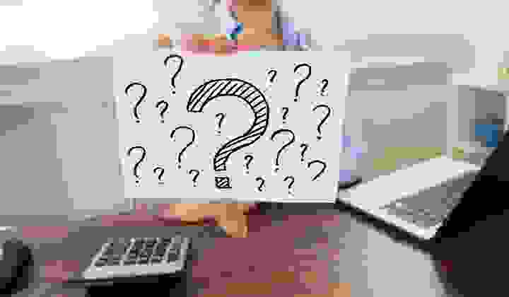 Woman at her desk with a calculator and laptop holding up a sign with lots of question marks