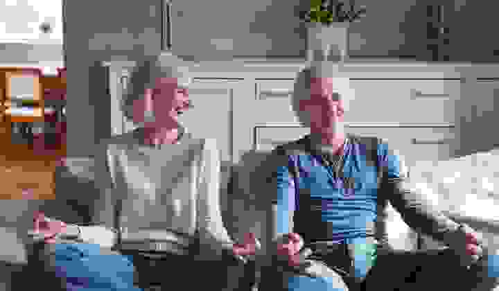 Mature couple sitting cross legged on couch smiling and doing the ohm finger pose