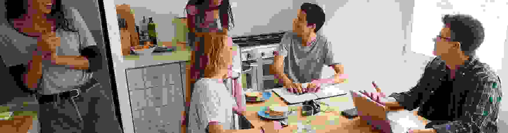 A group of laughing students gathered in a kitchen with laptops and sandwiches