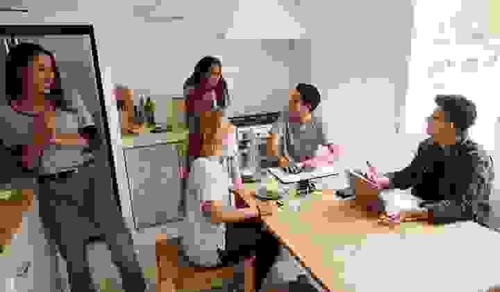 A group of laughing students gathered in a kitchen with laptops and sandwiches