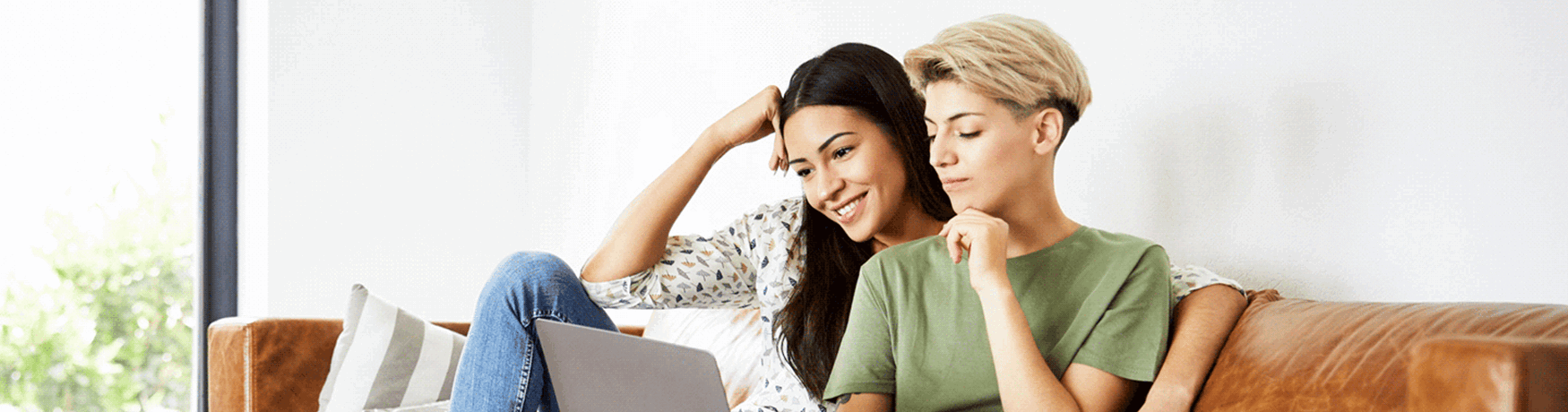 Two young women smiling on a couch, looking at a laptop