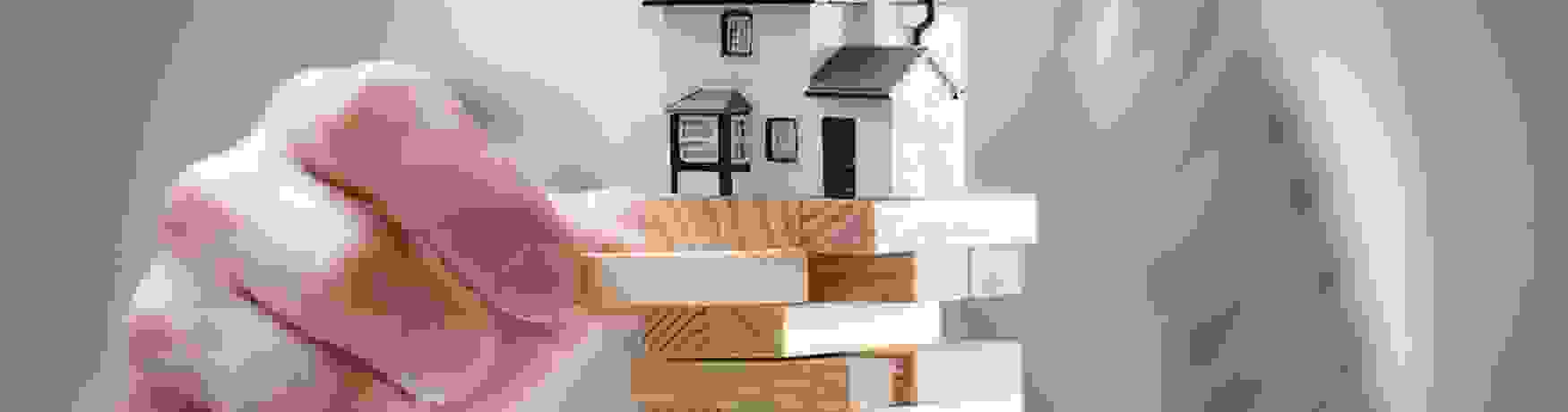 Sharp focus on a Jenga stack with a house on top as a man pulls a brick from the tower creating a huge risk