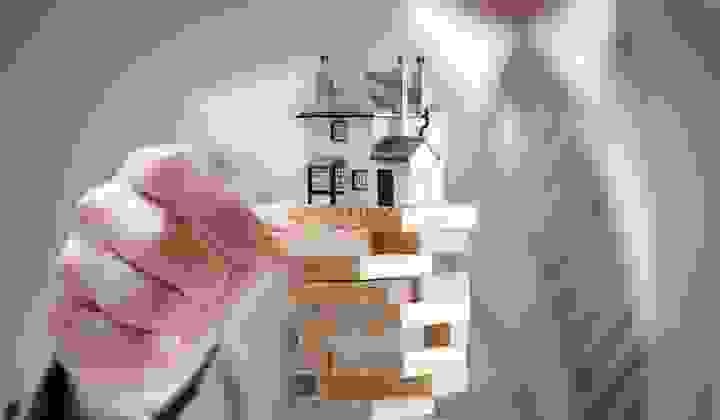 Sharp focus on a Jenga stack with a house on top as a man pulls a brick from the tower creating a huge risk