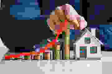 Growing stacks of coins with an arrow depicting inflation leads to a house with a man's hand holding a coin above a house model.