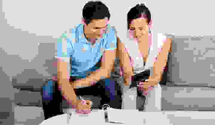 A young couple sit on their couch with a calculator and folder discussing their finances