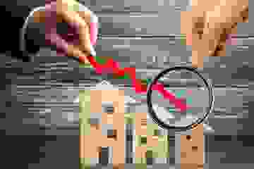 Magnifying glass over a red downward-pointing arrow and model houses showing the decrease in house prices