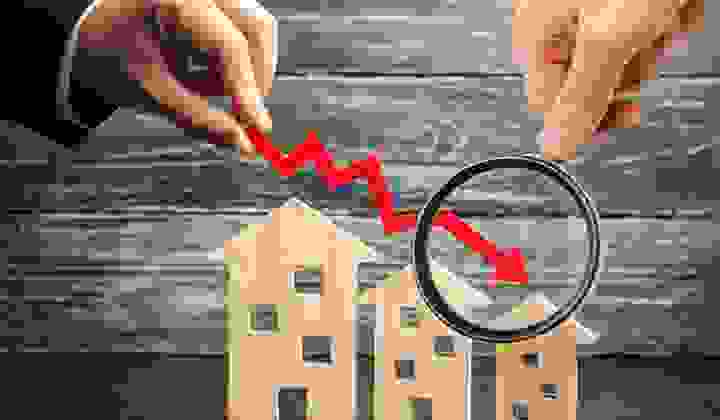Magnifying glass over a red downward-pointing arrow and model houses showing the decrease in house prices