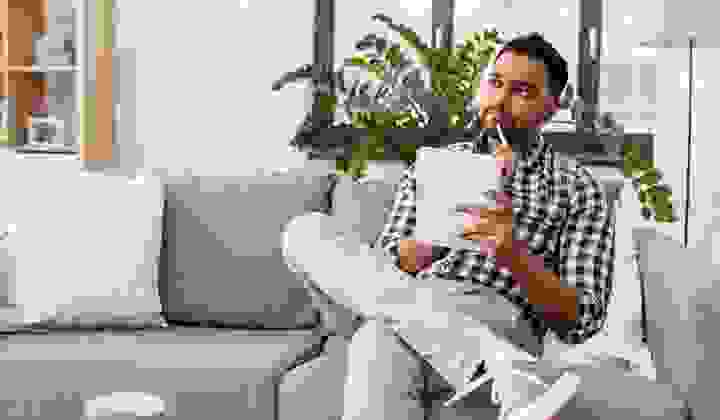 Young man sitting on his couch with pen and paper looking thoughtful