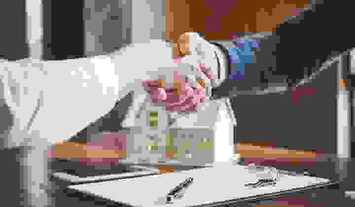 Close up of two people shaking hands over a real estate contract and set of keys with a house model in the background