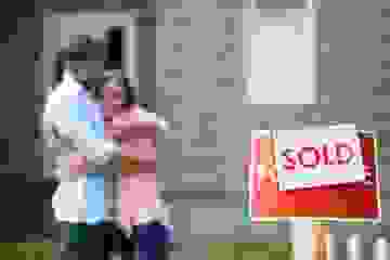 Couple hugs in the background in soft focus and a Sold sign is in focus in the foreground in front of a house