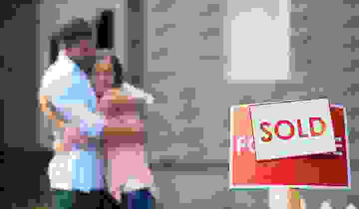Couple hugs in the background in soft focus and a Sold sign is in focus in the foreground in front of a house