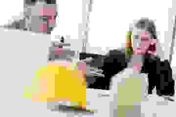 Business man discusses investment options with a woman in a suit in an office with a construction hat and house model blurred in foreground