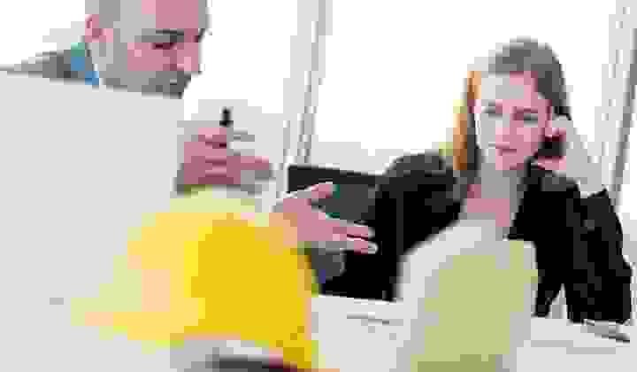 Business man discusses investment options with a woman in a suit in an office with a construction hat and house model blurred in foreground