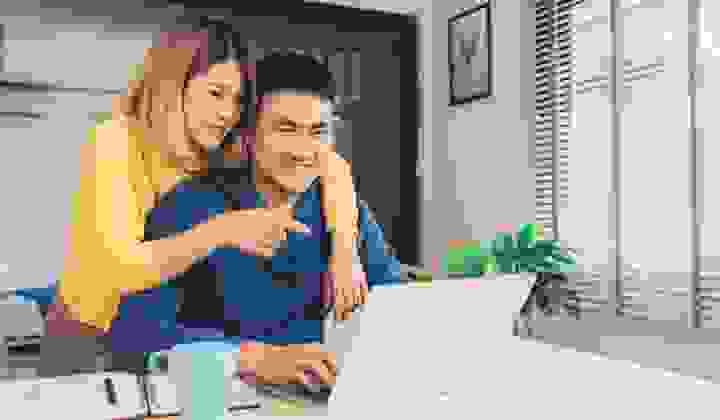 A smiling young couple look at their real estate listings on their laptop at home